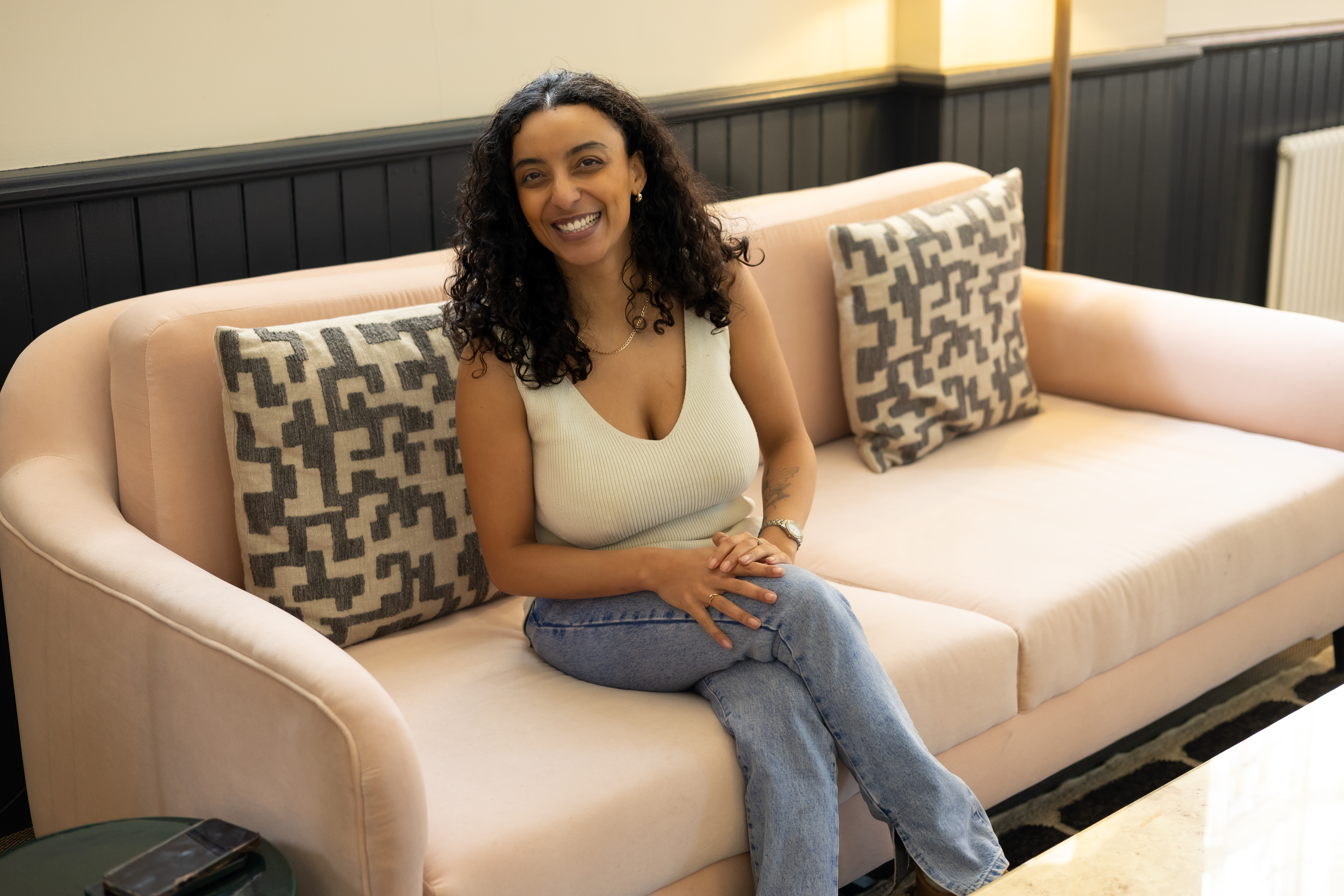 Simone sits on a pink couch smiling. She is wearing jeans and a white knit tank top. She has shoulder length curly brown hair and light brown skin.